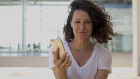 Smiling-brunette-woman-holding-smartphone-and-looking-at-camera